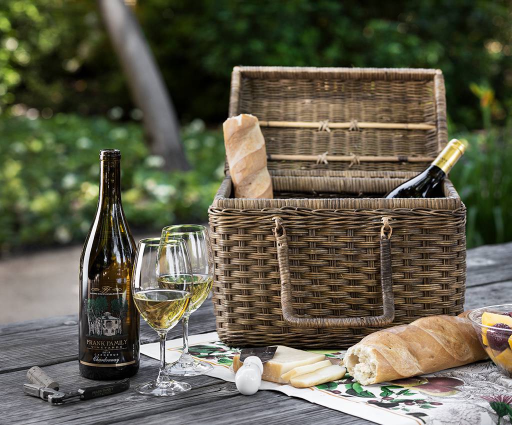 A picnic basket sits on a table with food and wine beside it