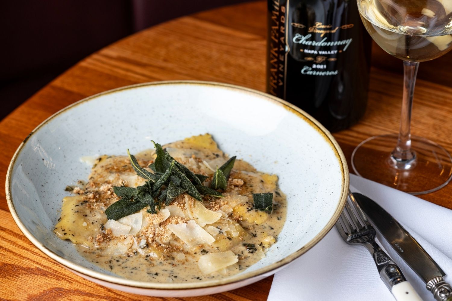 A bowl of creamy butternut squash ravioli topped with crispy sage alongside a glass of Lewis Vineyard Chardonnay.