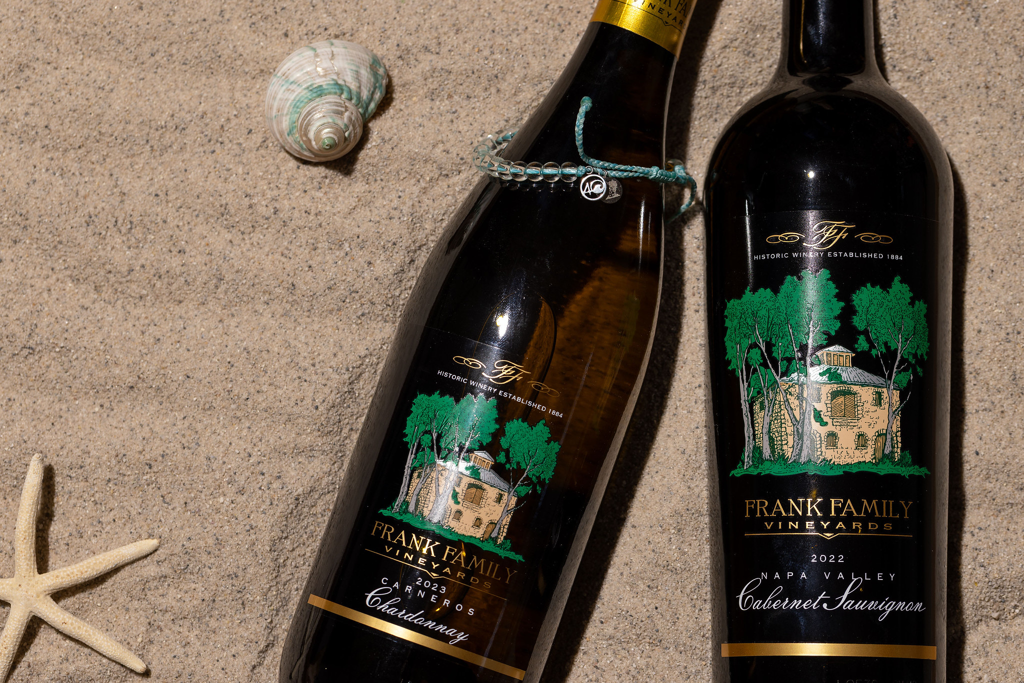A bottle of Carneros Chardonnay on the beach alongside a black 4ocean x Frank Family custom bracelet and bag