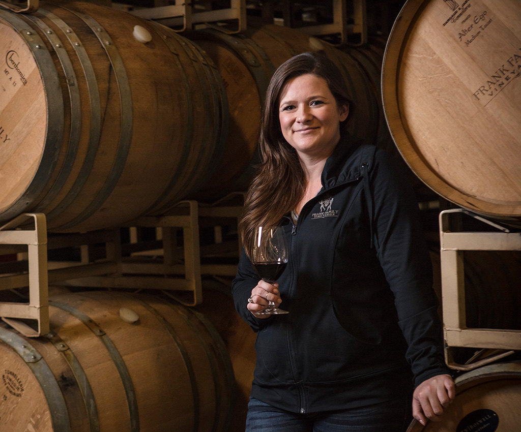 Corey poses with a glass of wine in hand in front of wine barrels