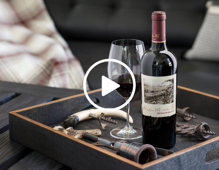 A bottle and glass of wine sitting atop a wood table with a plaid blanket draped in the background.