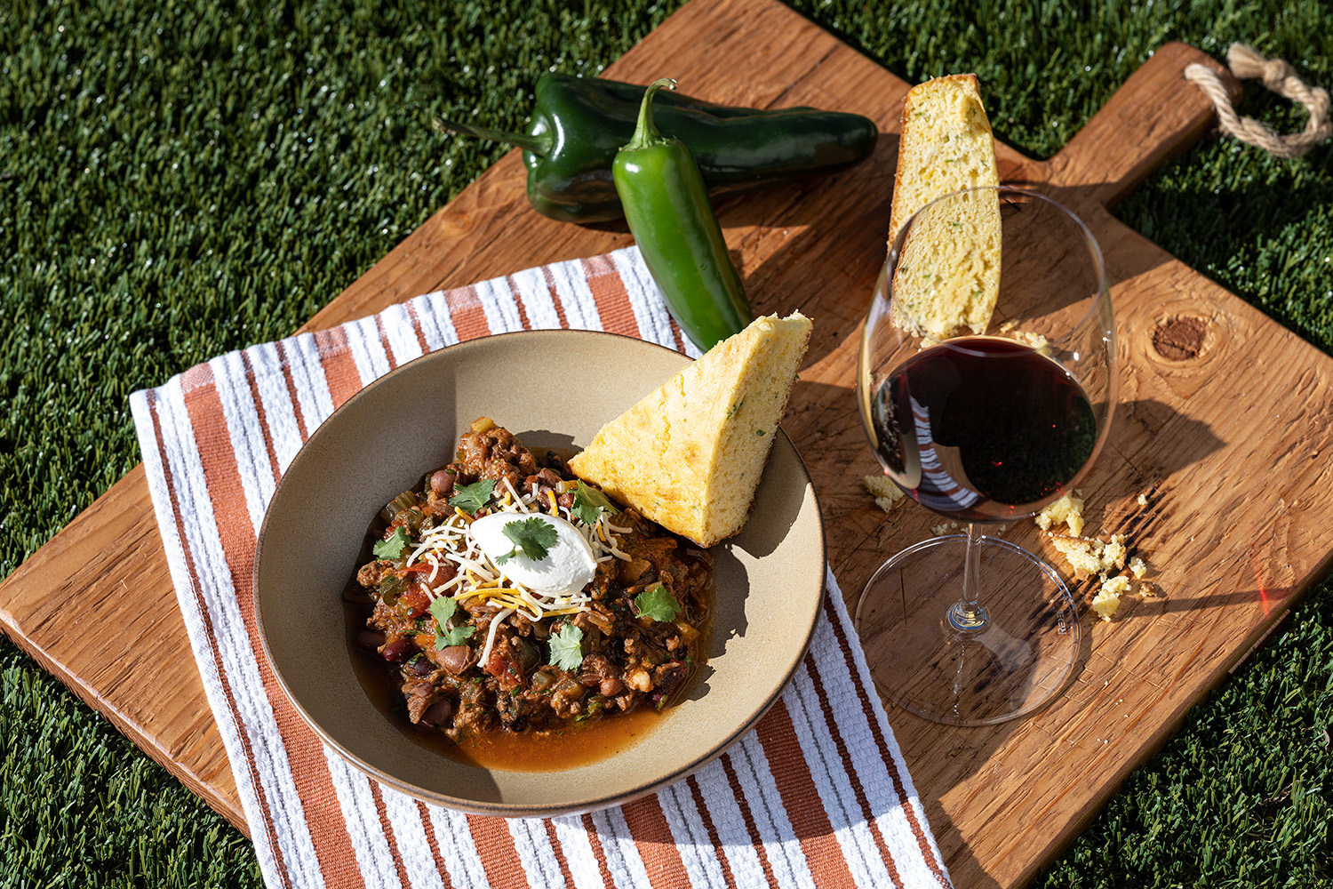 A bowl of chili on a cutting board next to cornbread, jalapeños, and a glass of red wine